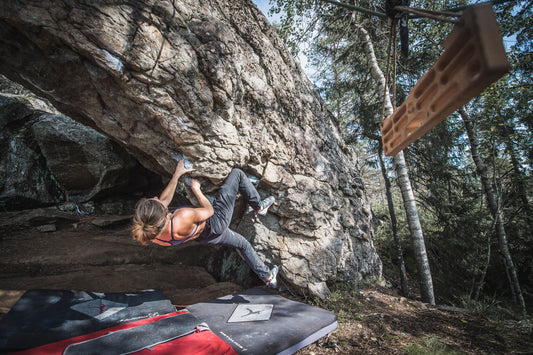 FIKA Vetelängd portable hangboard hanging at a bouldering crag with a climber in the background.